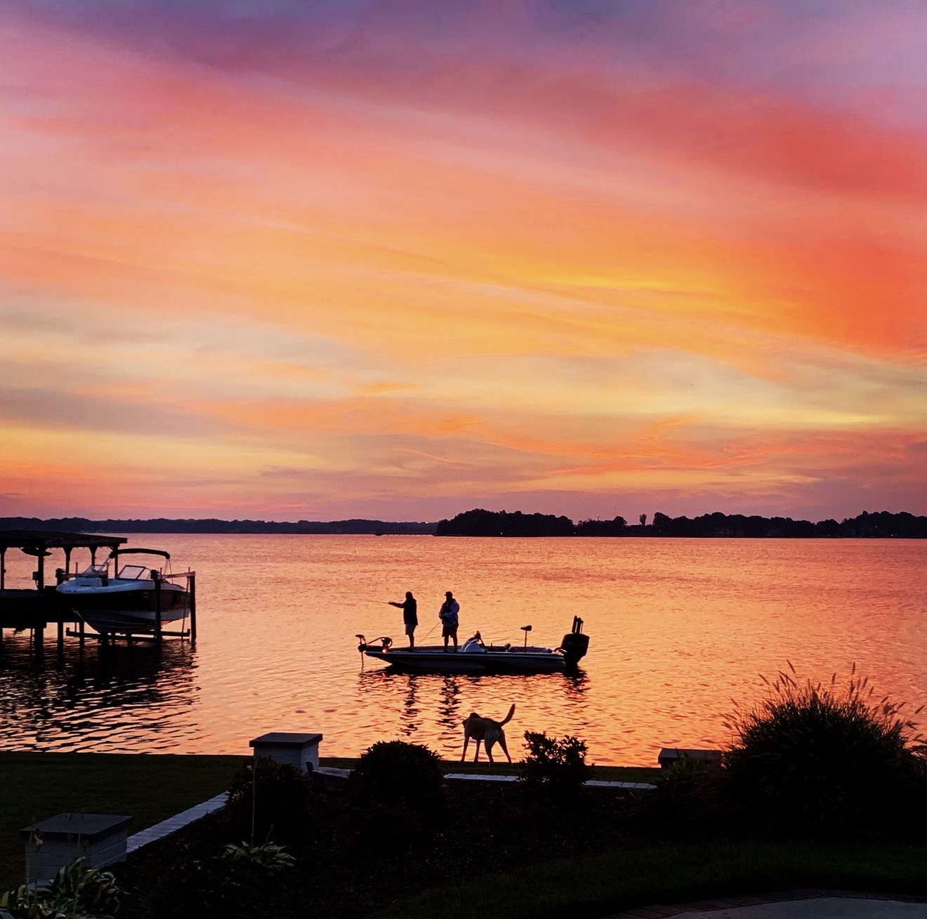 Two people fishing from a boat on Lake Norman at sunset while a dog watches from the shore